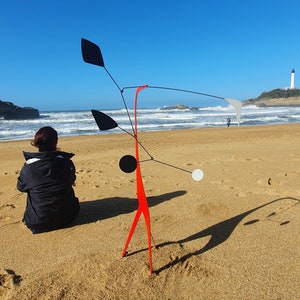 May include: A red metal mobile sculpture with black and white circular shapes and black leaf-shaped forms hangs on a beach. A person sits on the sand in front of the sculpture.