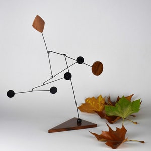 May include: A black metal mobile sculpture with wooden geometric shapes hangs from a wooden triangular base. The mobile features black circles and wooden squares and diamonds.