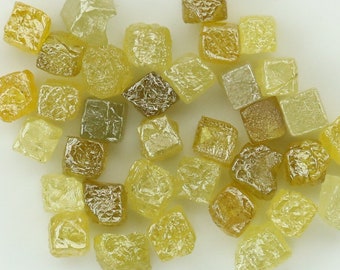 Puede incluir: Una colección de pequeños diamantes en bruto, amarillos y marrones. Los diamantes son de varias formas y tamaños, y están dispersos sobre una superficie blanca.