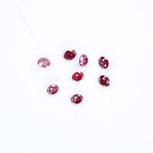 May include: A collection of eight oval-shaped, pink and red gemstones. The gemstones are scattered on a white surface.