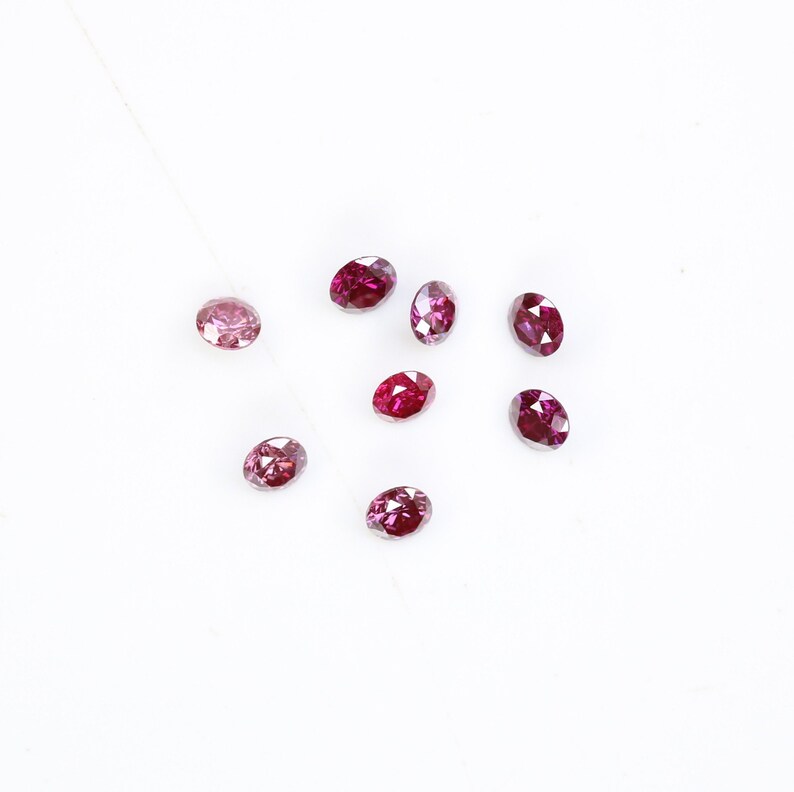 May include: A collection of eight oval-shaped, pink and red gemstones. The gemstones are scattered on a white surface.