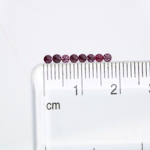 May include: A row of ten small, round, purple gemstones lined up on a white surface next to a ruler marked in centimeters.