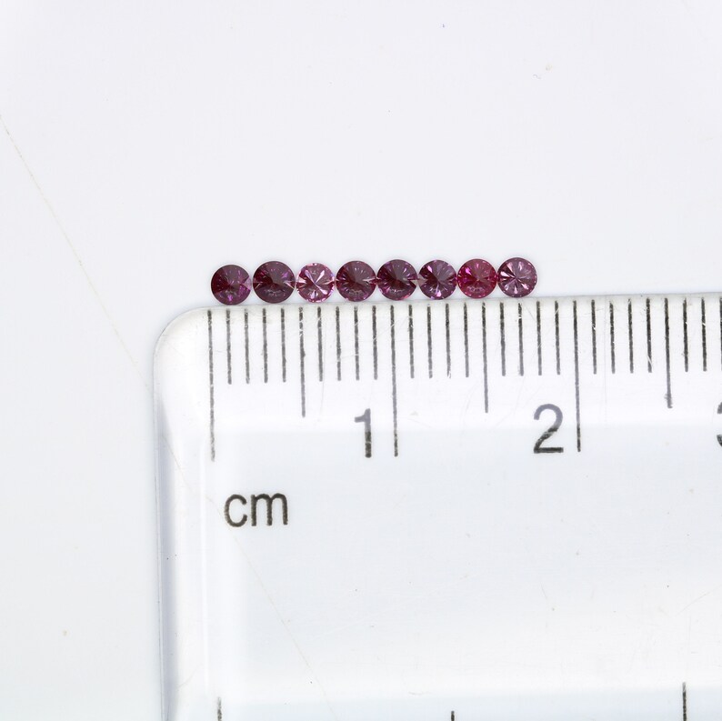 May include: A row of ten small, round, purple gemstones lined up on a white surface next to a ruler marked in centimeters.