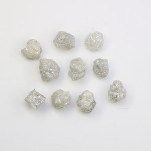 May include: A collection of ten rough, grey diamonds. The diamonds are various shapes and sizes, and they are all clustered together on a white surface.