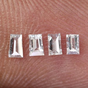 Puede incluir: Cuatro gemas claras de corte rectangular con una ligera forma trapezoidal. Las gemas se colocan sobre un fondo color piel, mostrando su claridad y facetas. Estas pequeñas piedras brillantes probablemente están destinadas a la fabricación de joyas.