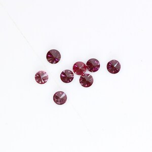 May include: A group of ten small, round, faceted, purple gemstones on a white background.