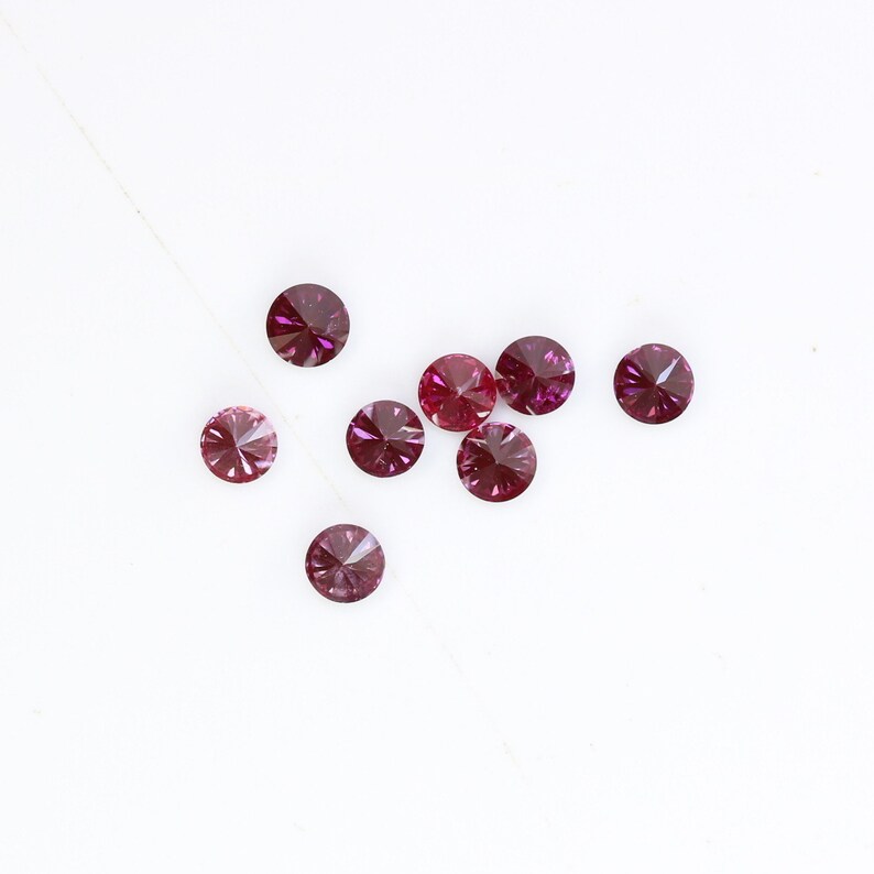 May include: A group of ten small, round, faceted, purple gemstones on a white background.