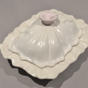 May include: A rectangular white ceramic butter dish with a ruffled edge base and a matching lid. The lid features a shell-like fluted design and is topped with a small, pale pink flower-shaped handle. The entire piece has a glossy finish.