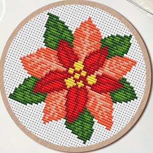 Puede incluir: Un patrón de punto de cruz de una flor de pascua en un aro de madera. La flor presenta pétalos rojos, naranjas y amarillos con hojas verdes. La palabra "PDF" está en un círculo blanco. También se ven tijeras y una aguja.
