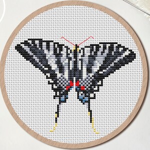 May include: A cross-stitch pattern of a butterfly with black, white, and gray wings, red accents, and yellow legs, displayed in a wooden embroidery hoop. The word "PDF" is in a white circle in the upper left corner. Scissors and a needle are also visible.