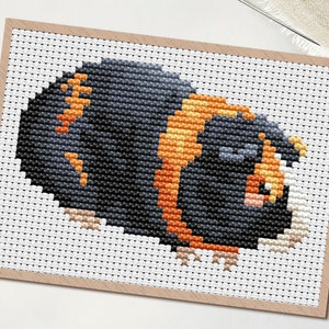 May include: A cross-stitch pattern of a guinea pig in black, orange, and tan. The image is on a white background with a wooden frame. The word "PDF" is in a white circle in the upper left corner. Scissors and a needle are also visible.