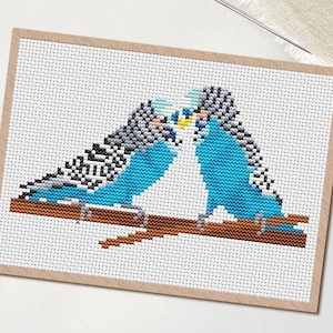 May include: Cross-stitch pattern featuring two blue parakeets perched on a brown branch, kissing. The birds have black and white patterned wings. The design is on white fabric, framed in a light brown border. The PDF logo is in the upper left corner.