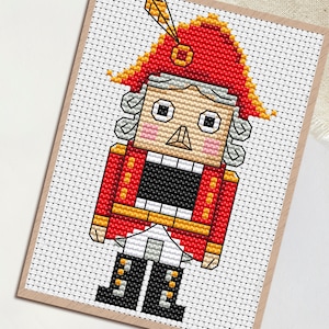 May include: A cross-stitch pattern of a Nutcracker soldier in a red coat and hat with gold accents. The design is on a white background with a grid pattern, and the word "PDF" is in a white circle in the upper left corner.