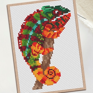 May include: A cross-stitch pattern PDF featuring a colorful chameleon perched on a branch. The chameleon is rendered in shades of red, orange, yellow, and green, with a curled tail. The design is displayed on a white grid background with a brown border.