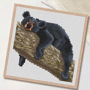 May include: Cross-stitch pattern of a black bear resting on a tree branch. The bear is depicted in shades of black and gray, with a brown branch. The design is on white fabric, framed in a light brown border. The word "PDF" is in a white circle.