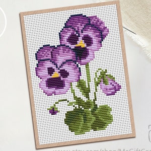 May include: Cross-stitch pattern of purple pansies with yellow centres and green leaves. The floral design is framed in a tan border. The image includes the text "PDF" in a white circle and a gift box icon.