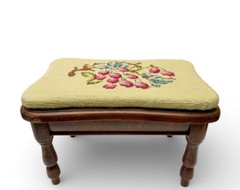 May include: A small, antique footstool with a needlepoint top. The cushion is a light yellow-green colour, embroidered with a cluster of pink grapes and green leaves. The wooden frame is a dark brown with turned legs.