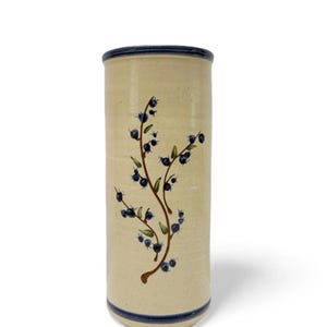 May include: A cylindrical ceramic vase with a speckled cream-colored finish. The vase features a hand-painted design of brown stems with small blue berries and green leaves. It has a dark blue band around the top rim and the bottom base.