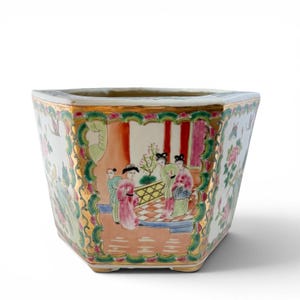 May include: A hexagonal porcelain planter with a gold rim and base. The planter features a hand-painted scene of figures in traditional attire, with floral and geometric patterns. The color palette includes pink, green, and gold accents.