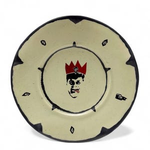 May include: A cream-colored ceramic plate with a black rim and a central illustration. The illustration features a face wearing a red crown. Black shapes are arranged around the plate's edge. The plate has a unique, artistic design.