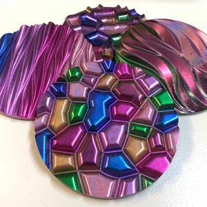 Puede incluir: Cuatro posavasos iridiscentes con texturas y diseños únicos. Un posavasos tiene un patrón ondulado, otro presenta un diseño geométrico de panal y los otros tienen superficies texturizadas. Los posavasos muestran una gama de colores, incluyendo azul, morado y verde.