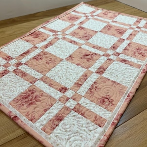 May include: A quilted table runner with a patchwork design. The runner features a pattern of alternating squares in white and a soft pink floral print. The edges are finished with a matching pink border. The dimensions are approximately 24 inches by 18 inches.