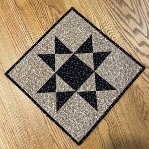 May include: A square quilted fabric mat featuring a central black star pattern with a delicate floral vine print. The star is surrounded by a light beige background fabric that also displays a matching intricate floral vine pattern throughout.