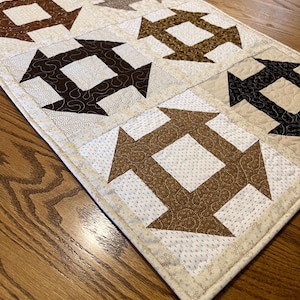 May include: A rectangular quilted table runner featuring a repeating block pattern of dark brown, tan, and cream fabric squares. Each block displays a central square surrounded by four arrow-like shapes, all finished with intricate machine quilting.