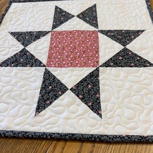 May include: A square quilted fabric mat featuring a central pink floral square surrounded by four black and white floral star points. The background is off-white fabric with a continuous meandering stipple stitch pattern and a dark floral border.