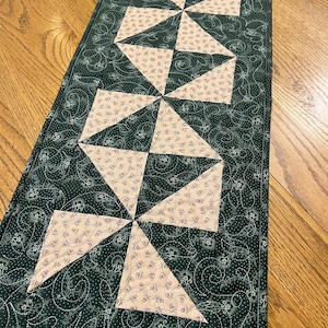 May include: A rectangular quilted table runner featuring a dark green fabric with a white swirling vine and floral pattern. The center displays a series of pinwheel blocks made from a light cream fabric with a small red and green floral print.