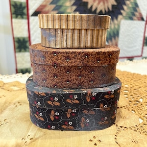 May include: A stack of three antique oval boxes. The top box has a striped pattern, the middle box has a floral pattern, and the bottom box has a floral pattern on a black background. The boxes are arranged on a lace tablecloth.