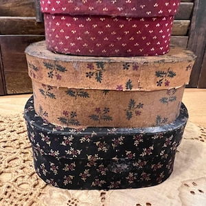 May include: Three antique oval storage boxes stacked on top of each other. The top box is a deep red with a small white floral pattern. The middle box is tan with a floral pattern. The bottom box is black with a floral pattern.