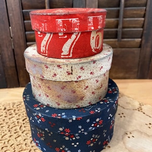 May include: Three stacked vintage round boxes. The top box is red with white musical notes and the number 76. The middle box has a floral pattern on a cream background. The bottom box is navy blue with a red and white floral pattern.