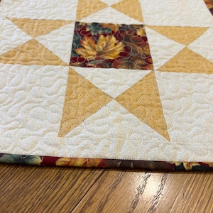 May include: A square quilted fabric mat featuring a central block with a dark brown background and a colorful autumn leaf print. The design includes a star pattern formed by light cream and golden yellow fabric pieces with stippled quilting.
