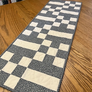 May include: A rectangular table runner with a geometric pattern. The runner features alternating blocks of cream and dark blue fabric. The dark blue fabric has a subtle floral print. The cream blocks have quilted details.