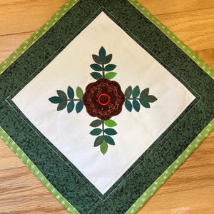 May include: A square, quilted table topper with a central embroidered rose design. The rose is a deep burgundy color, surrounded by green leaves. The background is white, framed by a dark green border and a light green patterned edge.