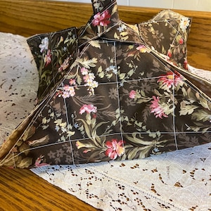 May include: A fabric bowl cozy featuring a dark brown background with a floral pattern of pink, red, and white flowers and green leaves. The item is quilted with a white grid stitch pattern and includes a fabric handle extending from the top center.