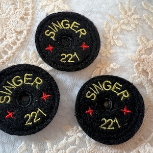 May include: Three black round Singer sewing machine bobbins with yellow embroidery. Each bobbin has a red star on either side and the text "Singer 221" in yellow embroidery.