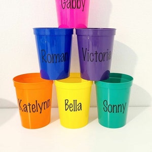 May include: Six colorful plastic cups with names written on them. The cups are pink, blue, purple, orange, yellow, and green.
