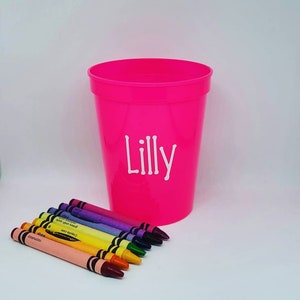 May include: A pink plastic cup with the name "Lilly" printed on it. The cup is filled with a set of eight crayons in various colors.