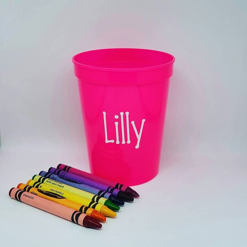 May include: A pink plastic cup with the name "Lilly" printed on it. The cup is filled with a set of eight crayons in various colors.