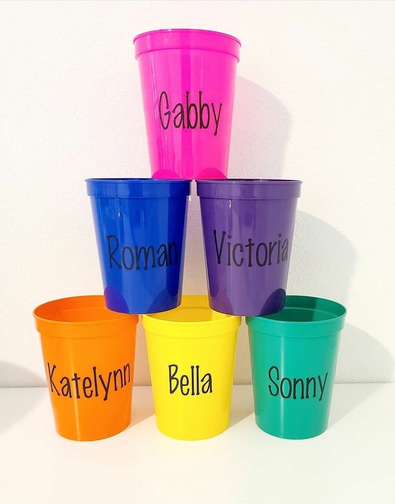 May include: Six colorful plastic cups with names written on them. The cups are pink, blue, purple, orange, yellow, and green.