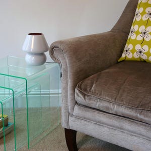 May include: Three clear glass nesting tables with a white ceramic lamp on the top table. The tables are in front of a brown armchair with a green and white floral patterned cushion.