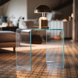 May include: A clear, U-shaped acrylic side table with a smooth, continuous surface. Resting on top of the table is a small, metallic gold lamp featuring a dome-shaped shade and a cylindrical base, connected by a thin, light-coloured power cord.