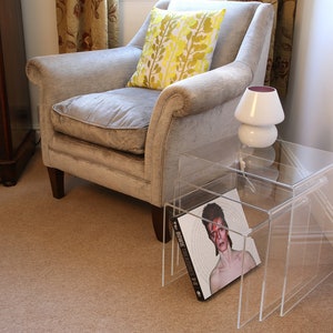 May include: A set of two clear acrylic nesting tables with a book on the top table. The book is titled "Bowie Treasures" and features a photo of David Bowie. A white lamp with a purple shade sits on the top table.