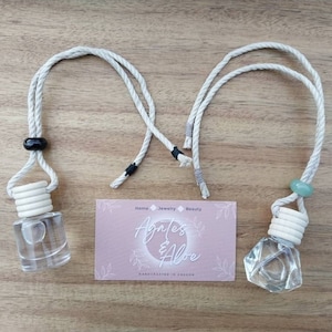 May include: Two car air fresheners with white rope hangers. The air fresheners are clear glass bottles with white stoppers. One bottle is round and the other is a geometric shape. The rope hangers have black and green beads. The image also includes a business card with the text "Agates & Aloe" and "Handcrafted in Oregon".