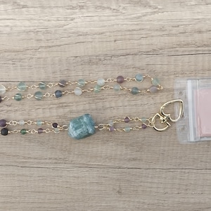 May include: A green and purple beaded lanyard with a gold chain and a large green stone charm. The lanyard has a gold heart-shaped clasp and is attached to a clear plastic badge holder with a pink card inside that says "Agates & Vibes".