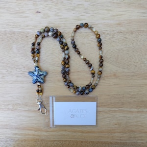 May include: A brown, gray, and gold beaded lanyard with a blue and white starfish charm and a clear plastic badge holder with the text "Agates & Aloe".