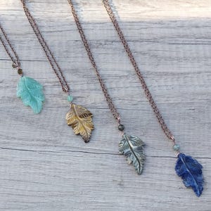 May include: Four necklaces with leaf-shaped pendants made of different types of stone. The necklaces are on a wooden surface. The leaves are carved from green, brown, gray, and blue stones.