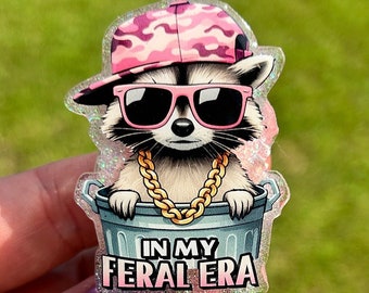 May include: A glittery badge holder featuring a cartoon raccoon wearing a pink camouflage hat, sunglasses, and a gold chain. The raccoon is sitting in a trash can with the text "In My Feral Era".